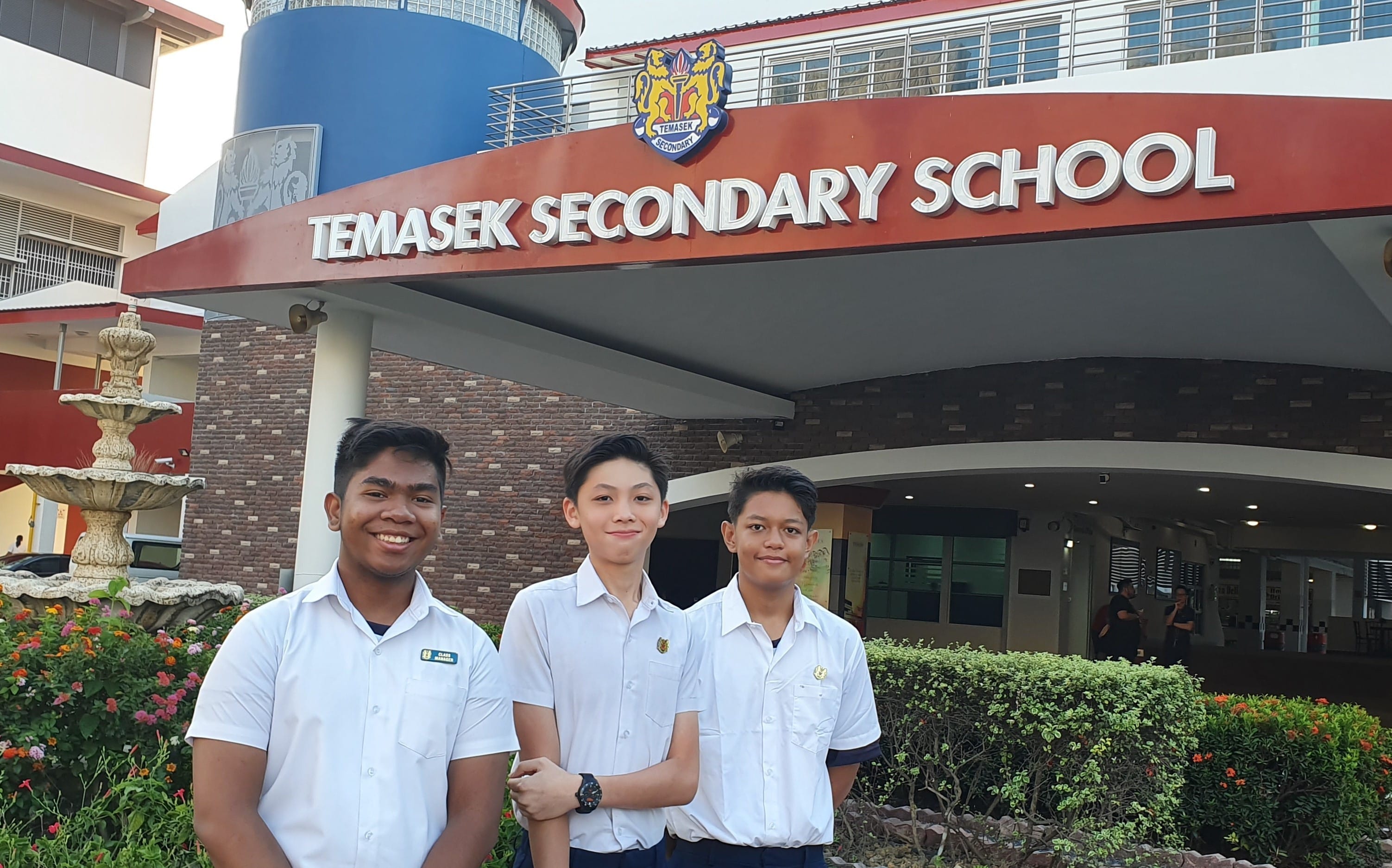 Three male students in white uniforms stand smiling in front of Temasek Secondary School entrance.